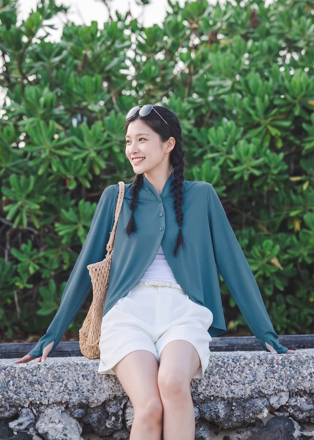 女模特兒穿著長袖抗UV防曬開襟罩衫與白色短褲,坐在綠意植物旁的石牆上休閒拍照