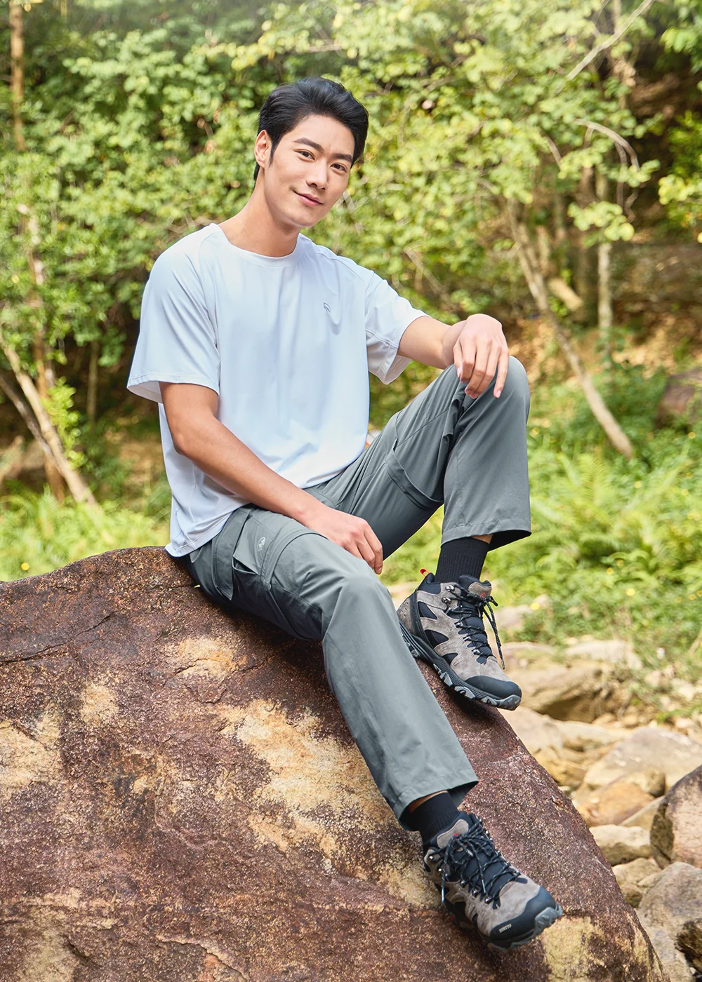男士中灰色抗UV-Suptex清涼彈性長褲穿搭。模特兒內搭白色短袖T恤,腳穿專業登山鞋坐在戶外岩石上。展現羽量輕彈布料的卓越靈活性,以及專為登山設計的立體膝褶與多口袋拉鍊口袋便利收納。
