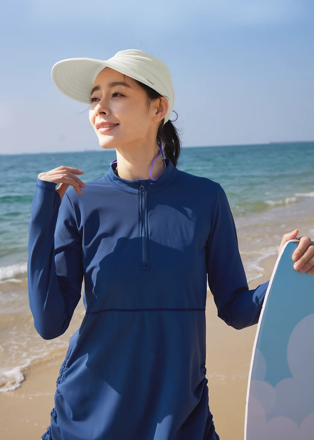 女款米白色涼感戲水彈性造型空心帽。模特兒站在海邊,配戴寬帽簷防曬空心帽,展示適合海邊戲水、輕鬆應對陽光與水域的運動時尚感設計。