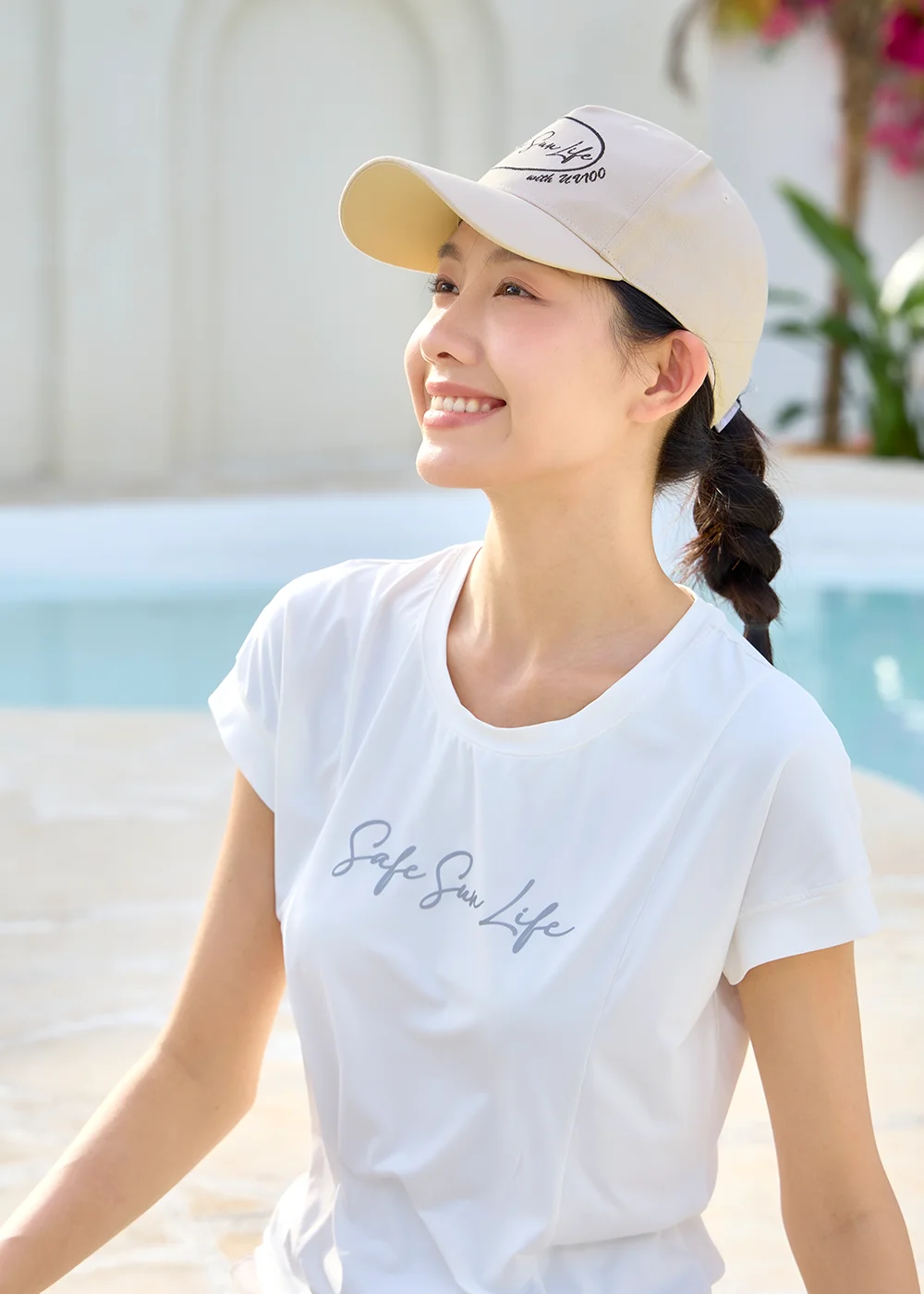 女性模特兒綁著單邊馬尾辮,身穿白色 Safe Sun Life 印花涼感 T 恤、頭戴米色經典款防曬老爹帽,在泳池畔側身仰望天空,展現簡約百搭的夏日抗 UV 遮陽帽穿搭。