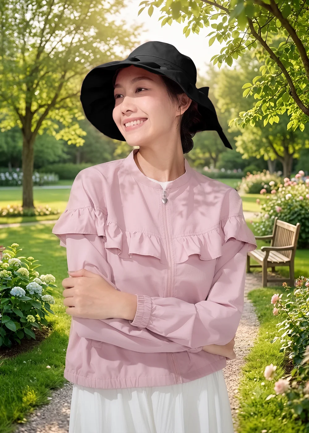 女性模特兒頭戴黑色 Aurelia 寬邊遮陽帽,身穿粉色荷葉邊防曬外套與白色長裙,雙手交疊走在開滿繡球花的英式花園步道旁,展現名媛質感的旅遊度假防風防曬穿搭。