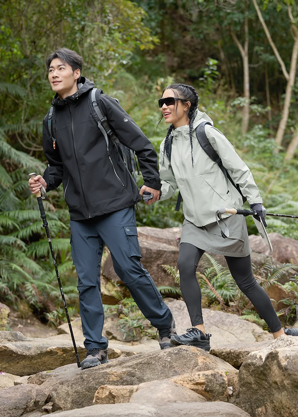男女模特兒手持登山杖結伴進行山林健行,男士身穿黑色防風連帽外套與藍灰色工裝長褲,女士身穿淺灰色防風外套與機能裙褲,展現工裝硬派的野外挑戰防護穿搭