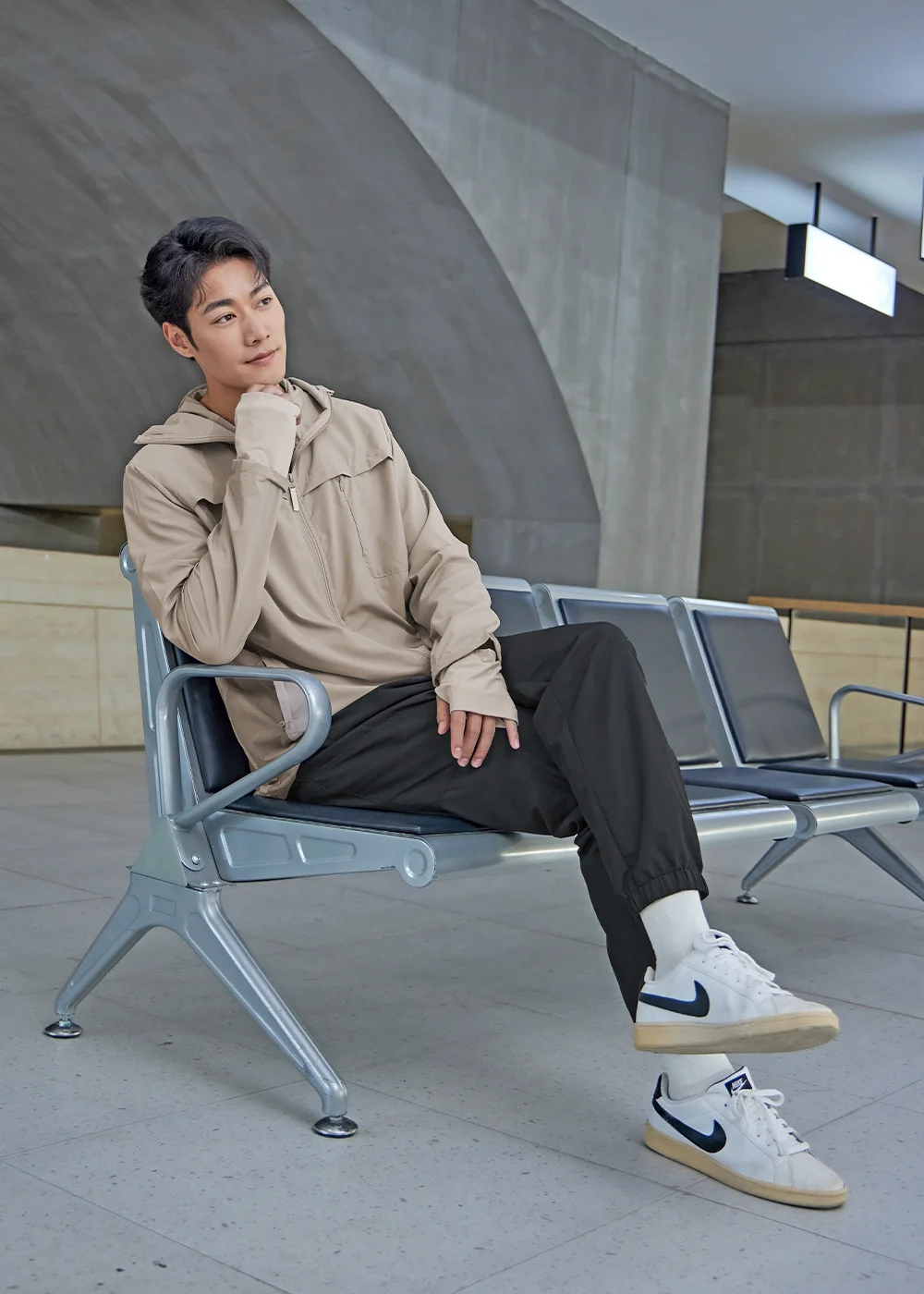 男模特兒身穿淺棕色防曬防風連帽外套與黑色Suptex九分防曬黑長褲,搭配白色運動鞋,舒適地坐在車站候車椅上思索,展現質感商務旅遊風格