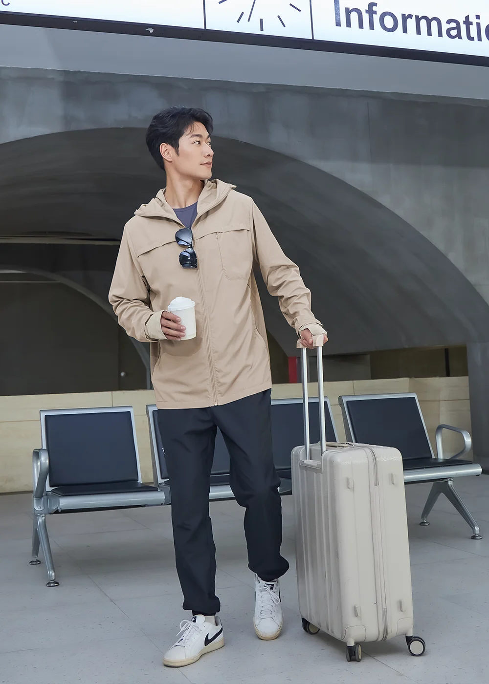 男模特兒手推行李箱、手拿咖啡杯,身穿防曬連帽外套與黑色九分機能長褲,在車站Information告示牌下漫步,營造舒適的機場商務穿搭