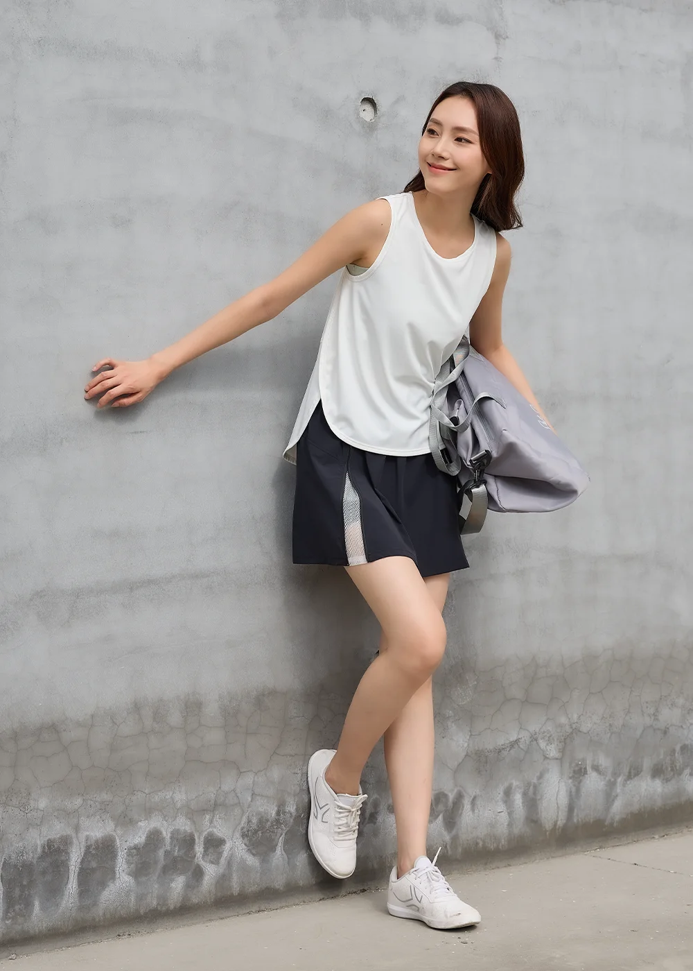 UV100 女生夏天涼感運動 OOTD:模特兒單手扶清水模牆面,身穿白色寬鬆運動背心、內搭運動內衣與黑色開叉運動短裙,展現透氣舒適的健身美學。