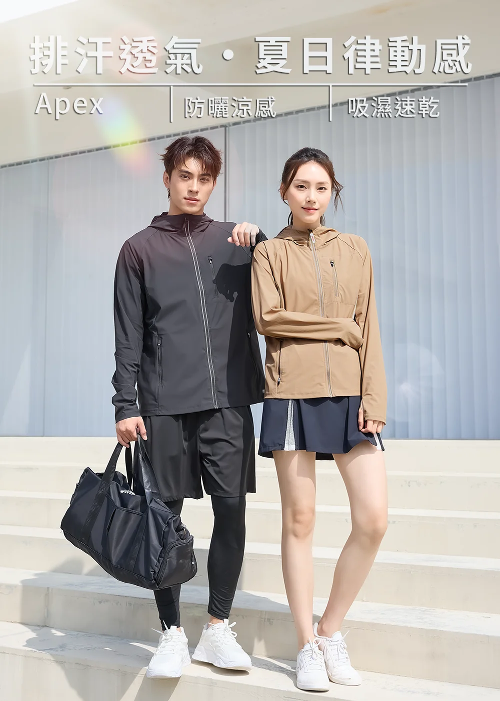 一對男女模特兒穿著機能連帽防曬外套站在白色階梯前。男模特兒穿著黑色外套搭配運動短褲與長內搭褲,手提黑色運動包;女模特兒穿著卡其色外套搭配深藍色運動短裙。