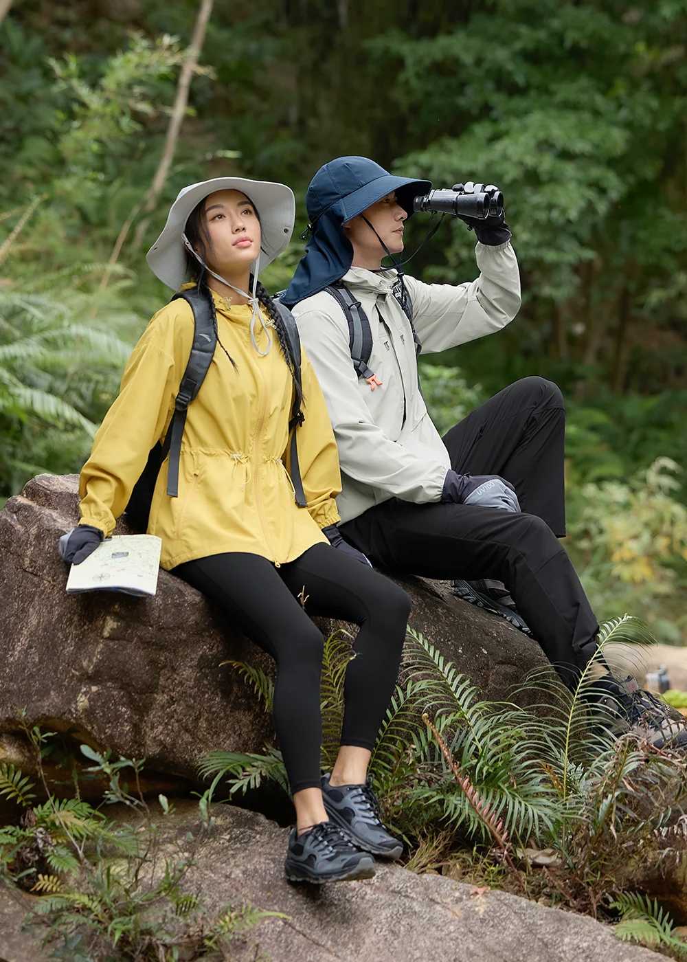 戶外專業登山防曬穿搭示範:男女模特兒佩戴寬簷防曬帽,搭配黃色與灰色連帽機能外套,於溪石景觀進行野外探索。