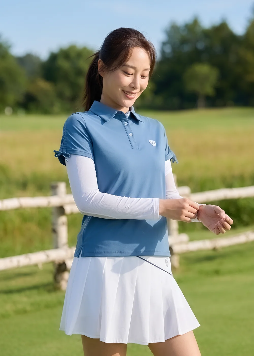 女模特兒綁馬尾微笑,身穿藍色短袖 Polo 衫、白色防曬百褶短裙與白色防曬袖套,在戶外木柵欄與草地背景前調整袖套細節。