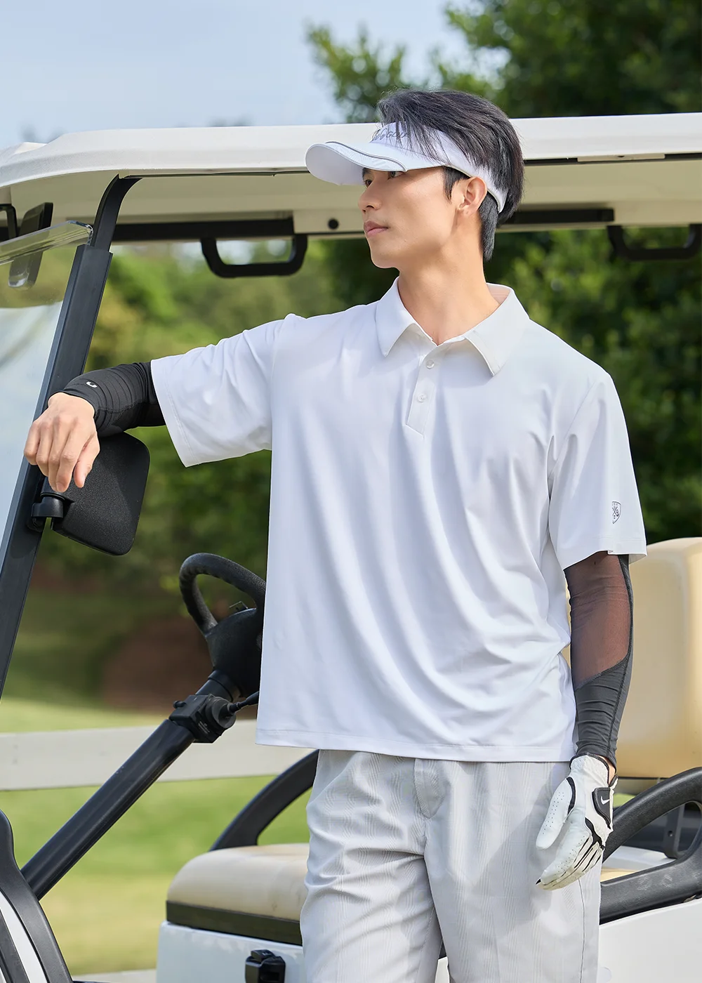 男模特兒戴白色運動遮陽帽、單手戴高爾夫球手套,身穿白色輕量 Polo 衫與黑色防曬袖套,側身扶著高爾夫球車展現運動 OOTD。