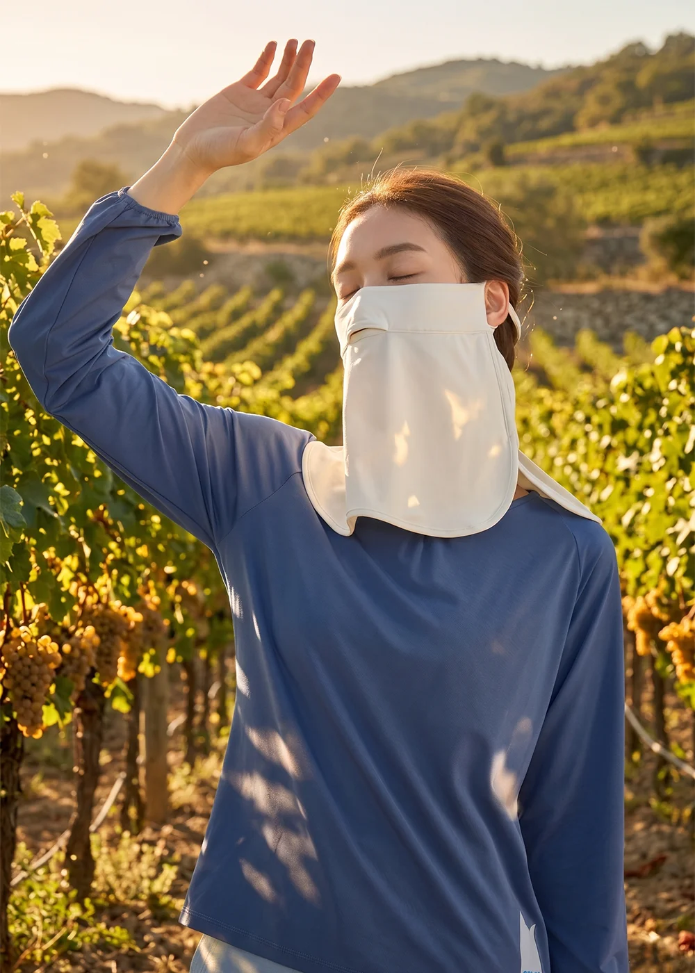 女性模特兒於陽光充足的戶外葡萄園配戴米白色UV100防曬涼感面罩,呈現夏日高效抗UV物理防曬情境