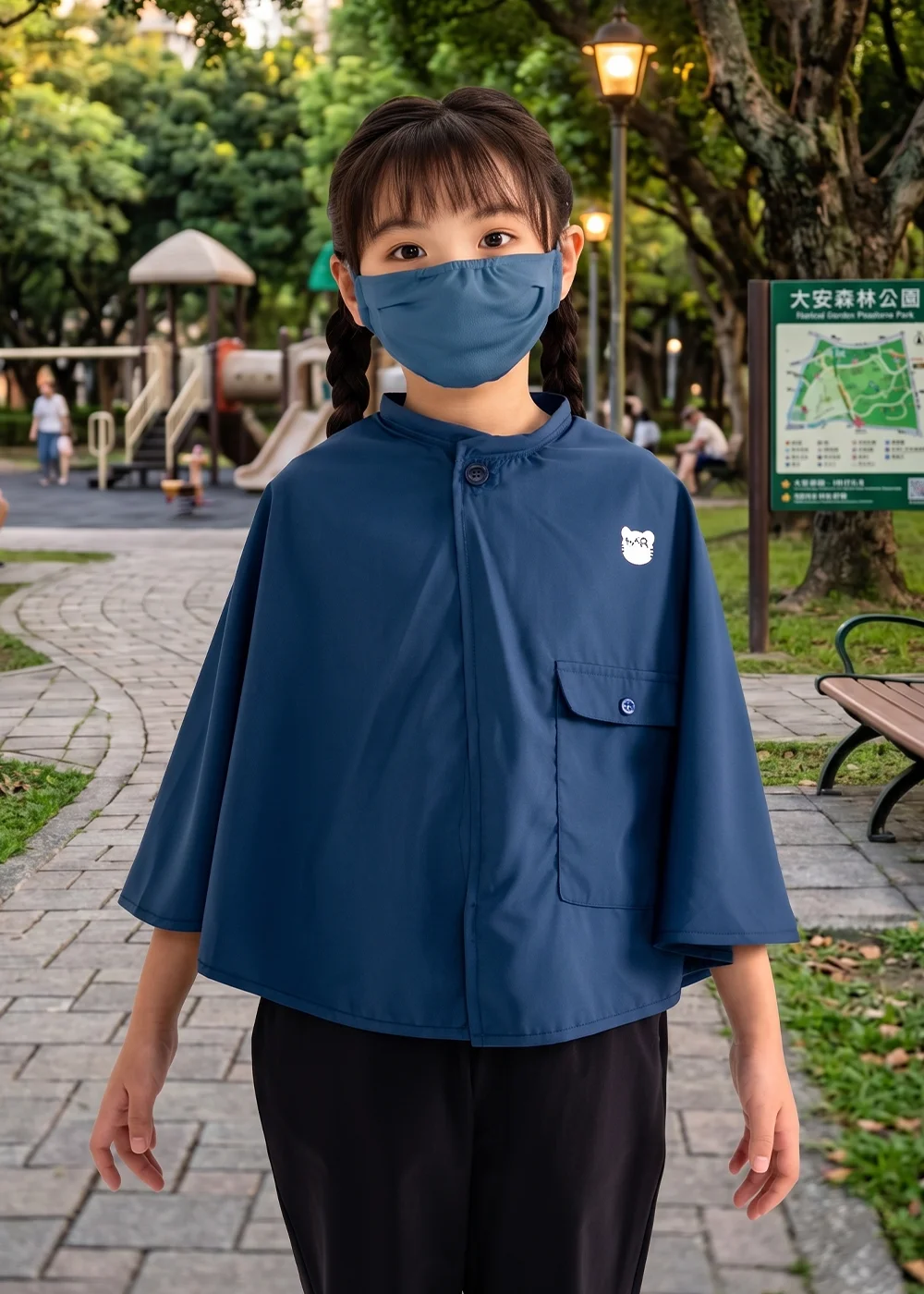 女童身著深藍色立領防曬斗篷搭配同色系口罩,置身於大安森林公園步道,胸前點綴簡約貓咪圖樣。