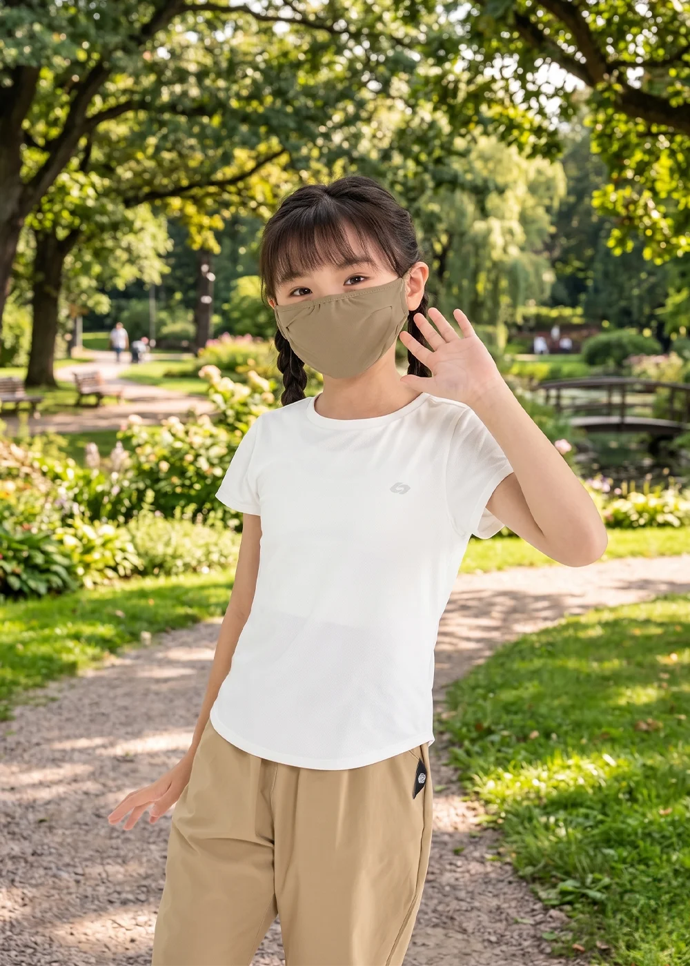 穿著純白排汗衫與卡其休閒褲的女孩,在綠意盎然的林蔭小徑向鏡頭揮手,配戴大地色調防曬面罩。