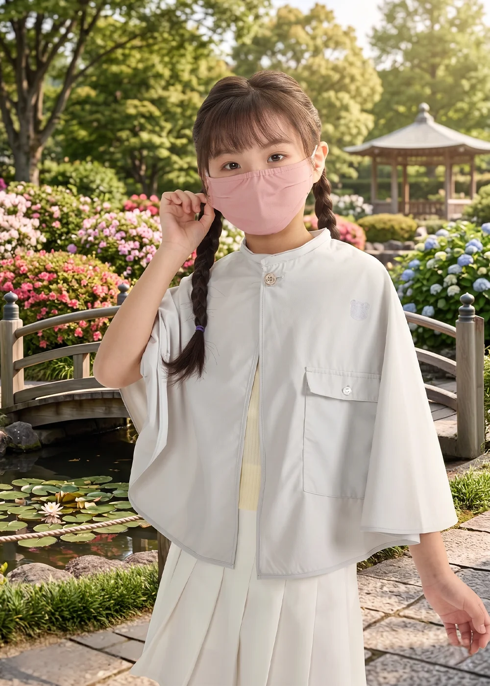 背景為日式庭園造景,孩童穿著淺灰色斗篷外套與粉色口罩,在小橋流水旁展現優雅與機能兼具的日常穿搭。
