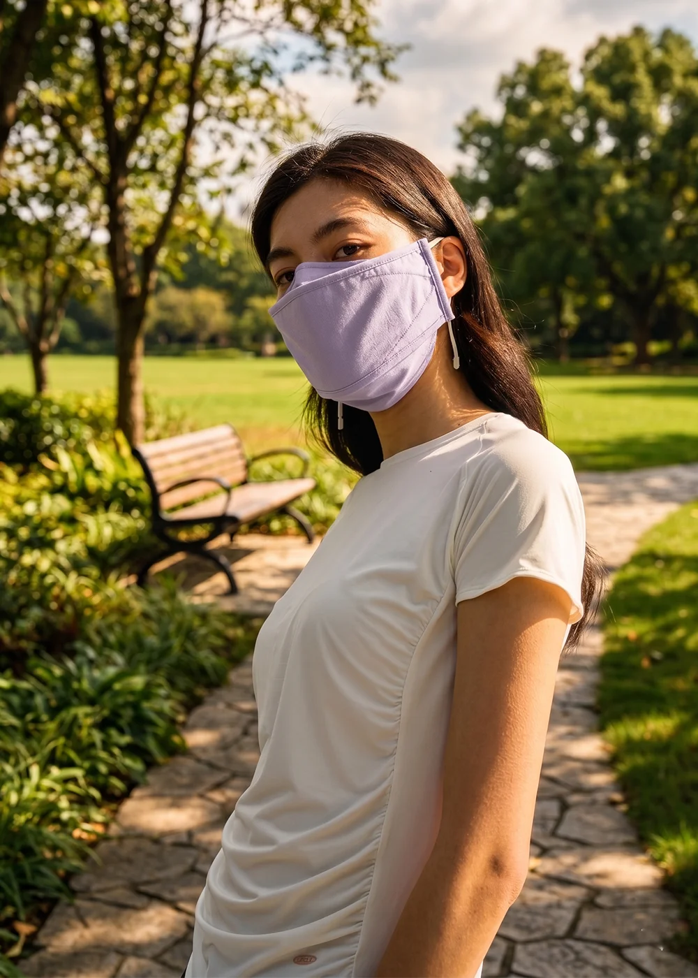 UV100 女生夏天透氣防曬口罩:模特兒身穿白色側邊抓皺休閒上衣,配戴粉紫色立體加大遮陽口罩,在陽光充足的公園綠地與木質長椅背景前展現戶外防曬 OOTD。