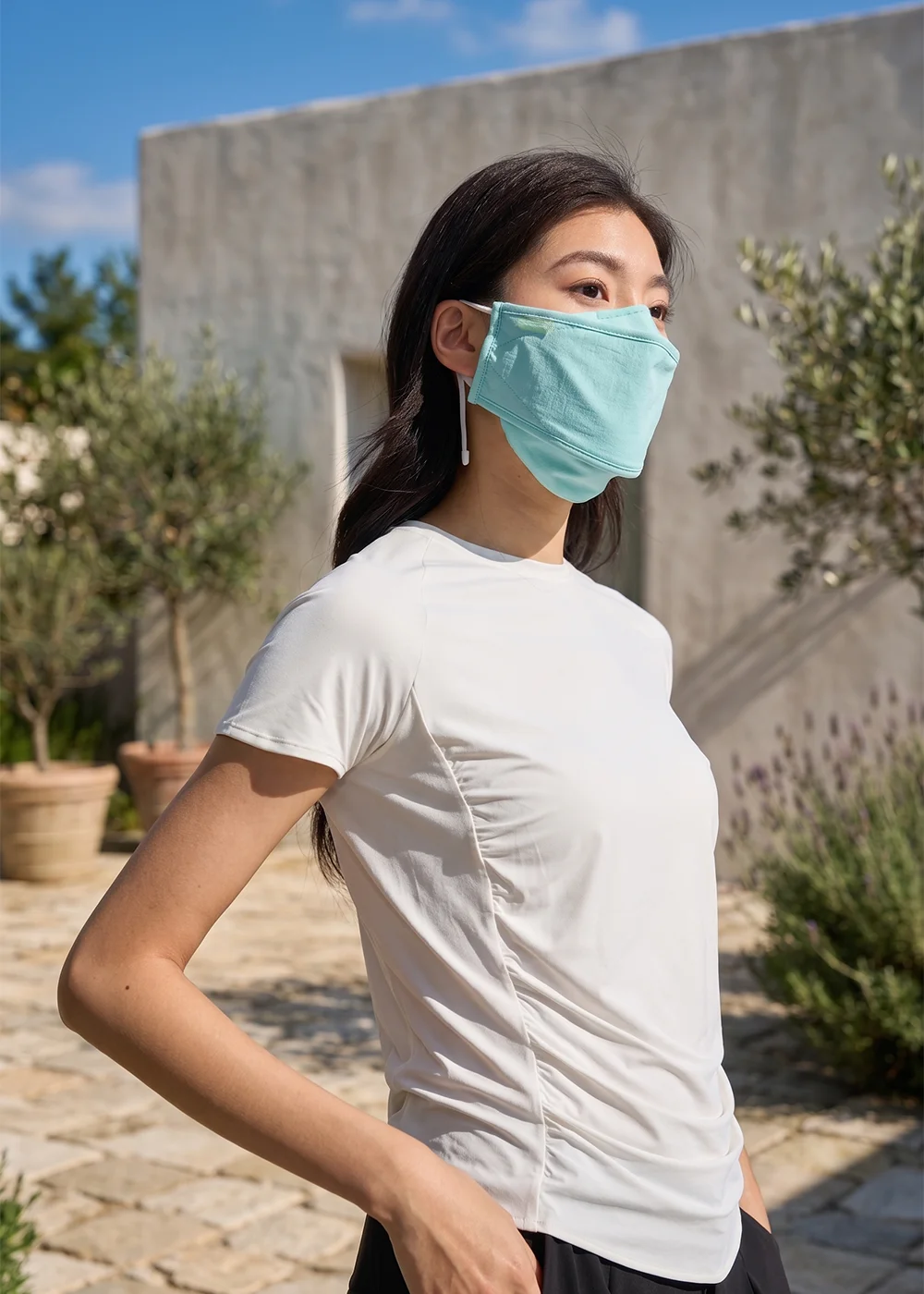 涼感機能防曬口罩推薦:女模特兒手插口袋,身穿白色排汗短袖上衣,配戴粉藍綠色加大立體防曬口罩,在歐風淺色建築與盆栽前展現清爽遮陽風格。