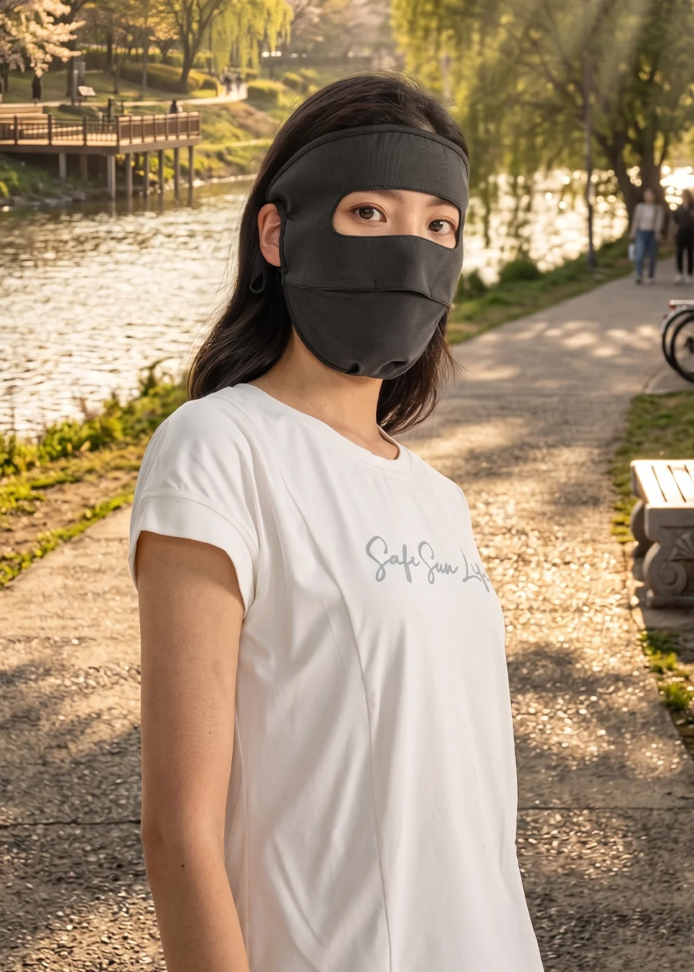 女模特兒穿著白色 T 恤佩戴黑色全臉防曬面罩,站在夕陽下的河濱步道,強調面罩在戶外休閒情境中的應用。