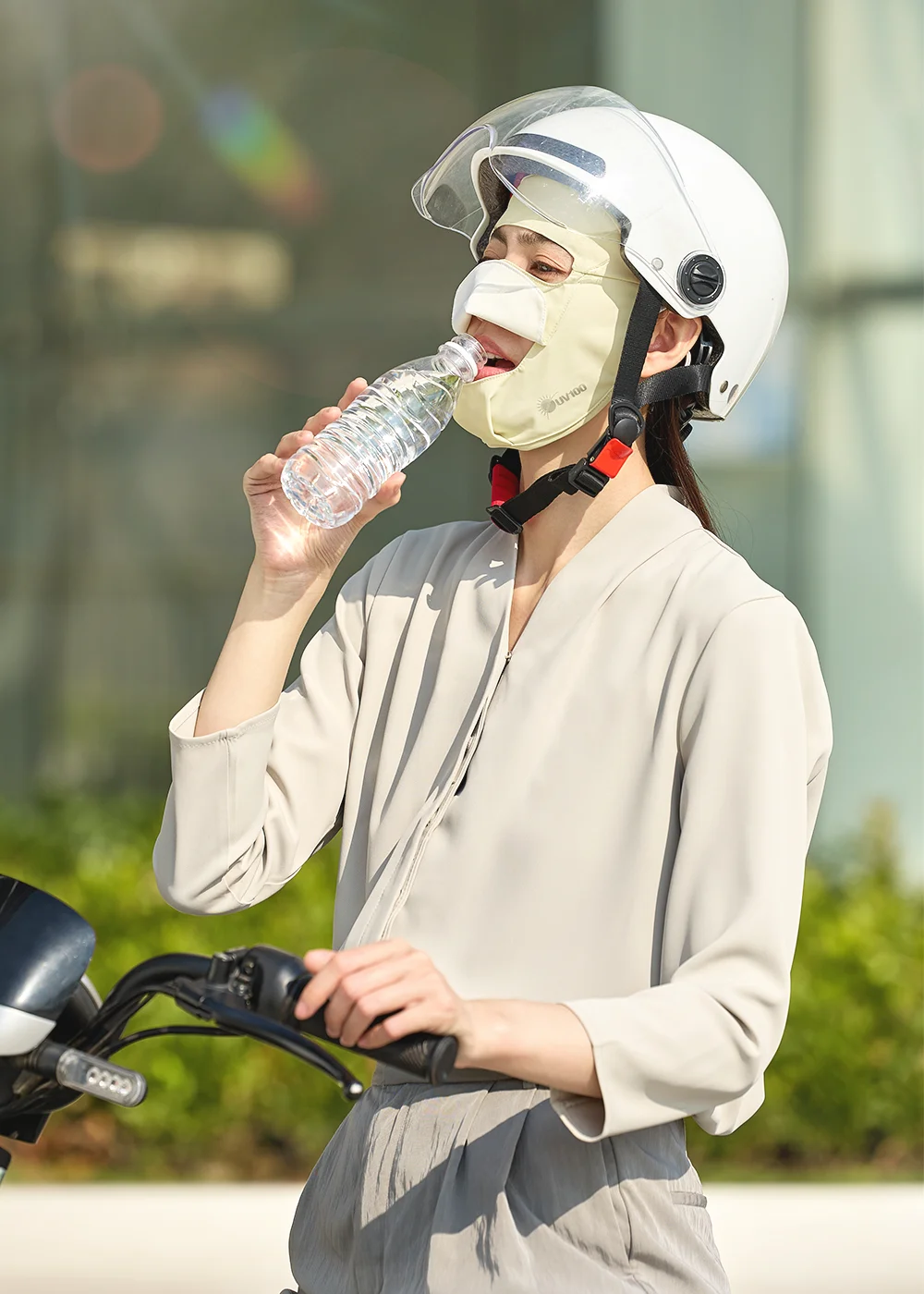 模特兒頭戴白色安全帽、騎乘機車時配戴 UV100 胚芽米款涼感排汗大開口透氣面罩,生動示範透過面罩下方的貼心大開口結構輕鬆喝水,展現騎車通勤時的便利機能。