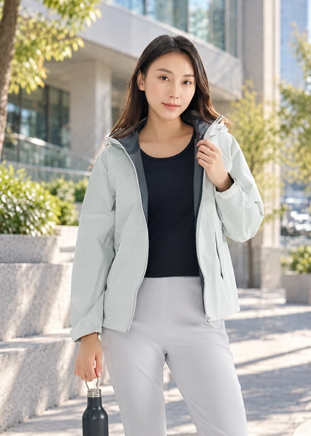 女性模特兒戶外穿搭展示:黑色科技羊毛背心外搭淺色連帽防風外套,手拿保溫瓶的半身照