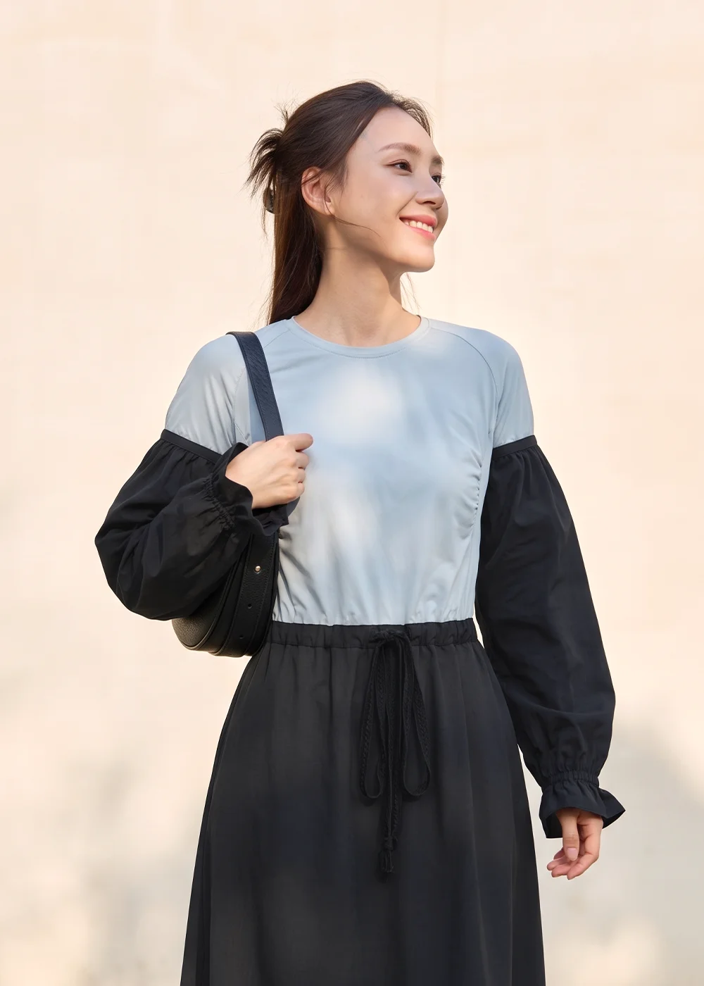 女模特兒身穿淺藍色排汗修身上衣,拼接黑色荷葉邊波浪防曬袖套,搭配黑色抽繩長裙與手提包,在簡約牆面背景前展現無懼烈日的時尚撞色機能穿搭