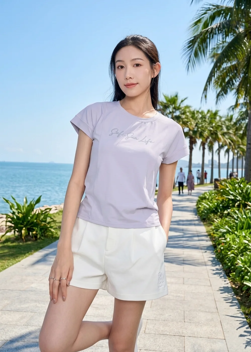 模特兒身穿淡紫色印花短 T 恤與白色短褲，站在陽光明媚的海濱棕櫚樹步道。