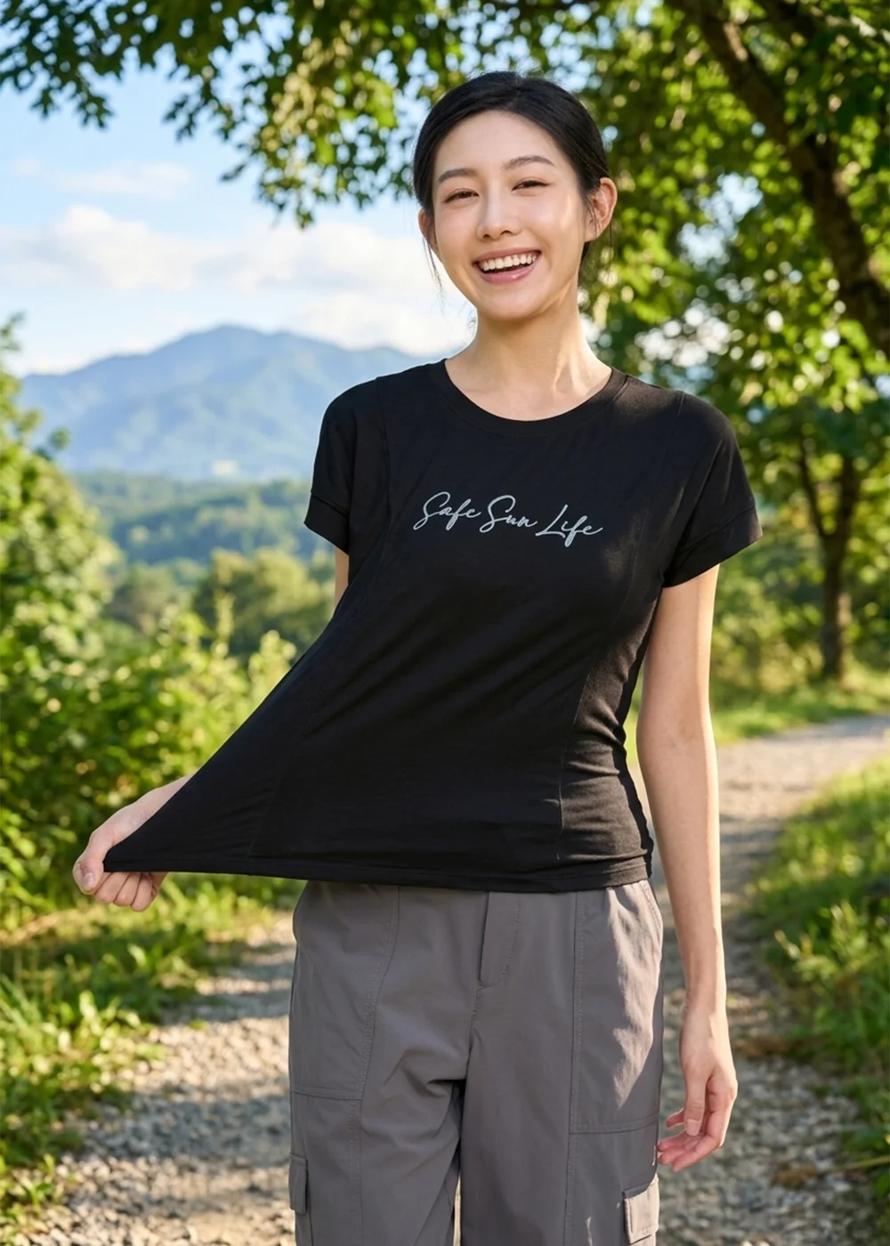 模特兒展示黑色彈性排汗衫，手拉衣服側邊呈現布料延展性，背景為綠意山徑。