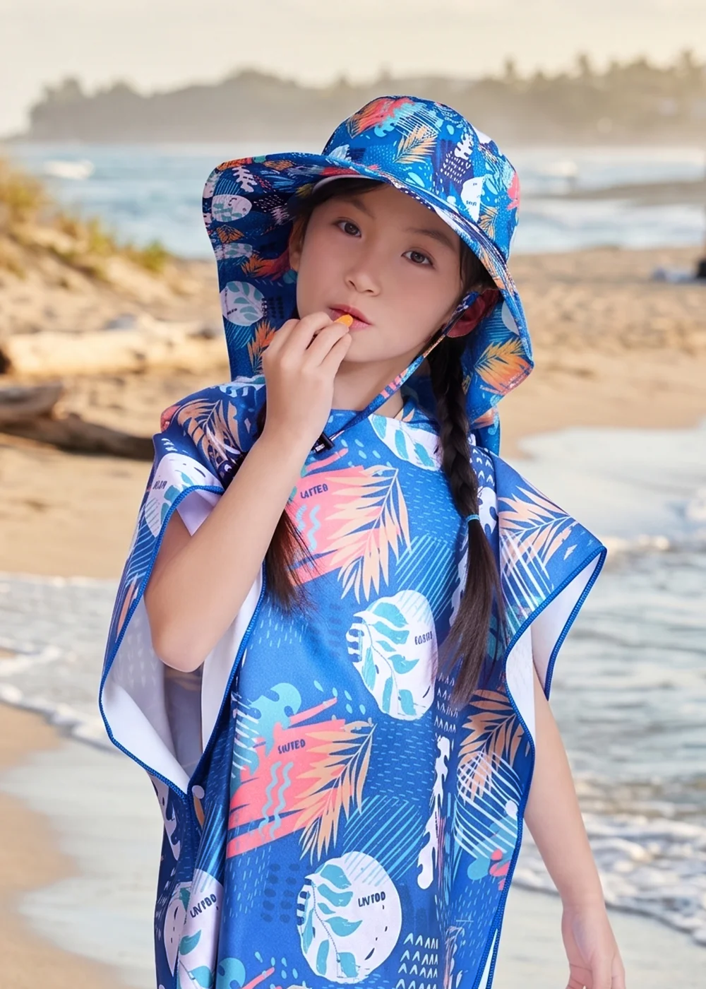 兒童防曬斗篷與防曬帽穿搭，小女孩戴著藍色熱帶葉片印花遮陽帽、身穿同款繽紛印花的防曬連帽斗篷披肩，在海灘背景前吃著點心，展示全方位兒童海邊防曬與抗紫外線保護。