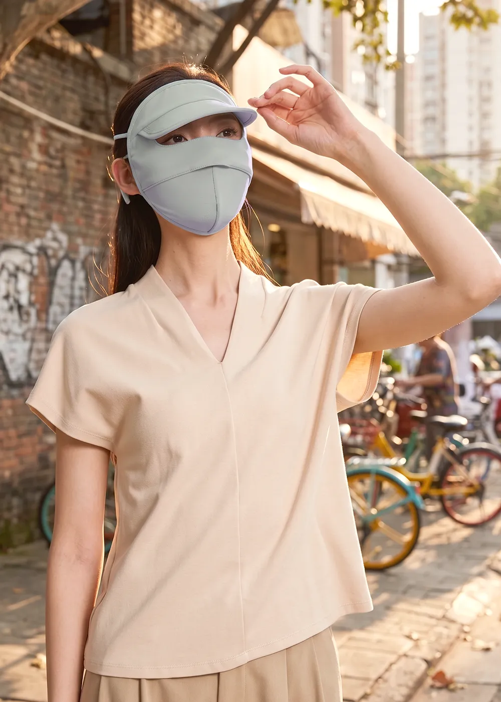女性模特兒穿著休閒V領上衣、配戴莫蘭迪綠涼感防曬面罩帽，在街頭與單車旁陽光下駐足