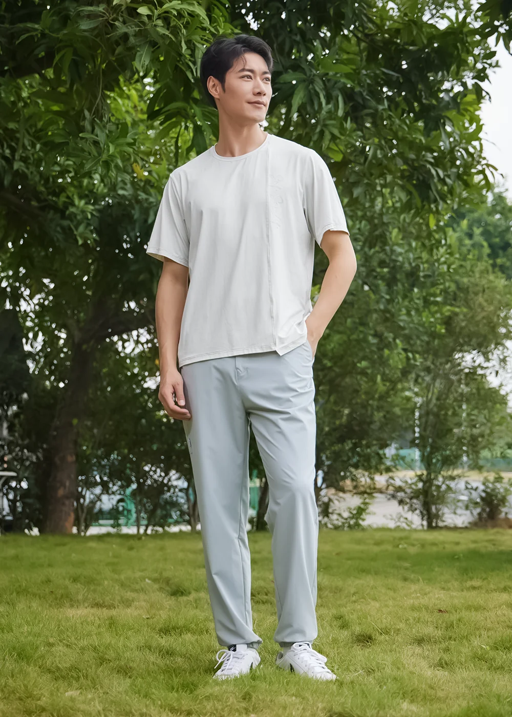 男模特兒身穿淺灰色機能上衣與淺藍色長褲，在草地綠蔭背景下展示全身休閒穿搭。
