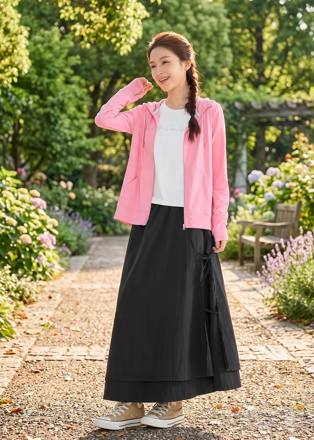 女生在花園中穿著 UV100 粉紅色連帽防曬外套，內搭白色 T 恤與黑色不對稱綁帶機能長裙，展現柔美與功能性兼具的戶外休閒穿搭。