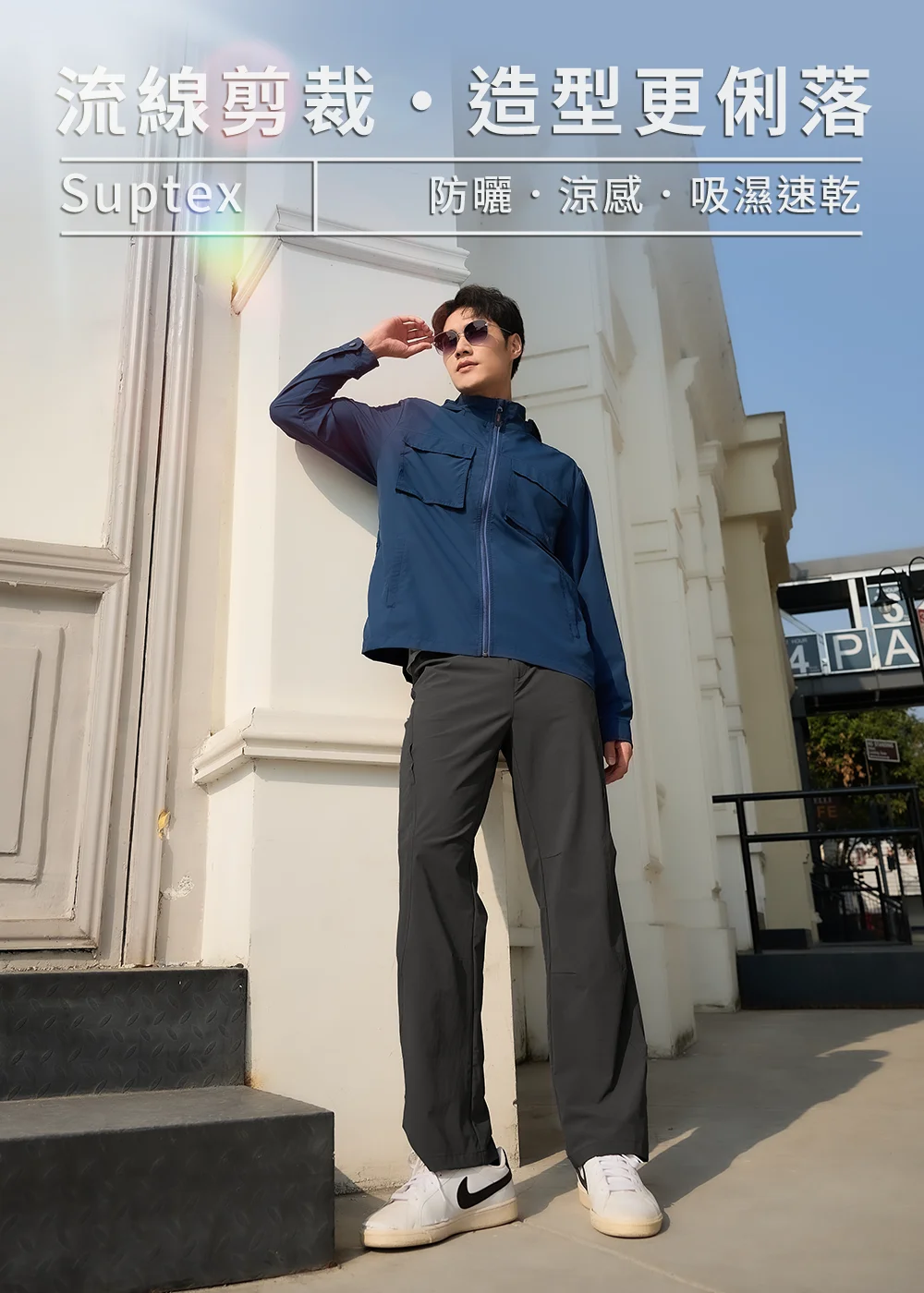 SUPTEX 防曬涼感吸濕速乾服裝宣傳圖。一名穿著深灰色長褲和藍色功能性外套的亞洲男子，佩戴墨鏡，斜靠在歐式白色建築門口，擺出時尚的姿勢。背景為藍天和遠處的建築。圖片上方有中文文案：‘流線剪裁 • 造型更俐落’和‘SUPTEX 防曬 • 涼感 • 吸濕速乾’。