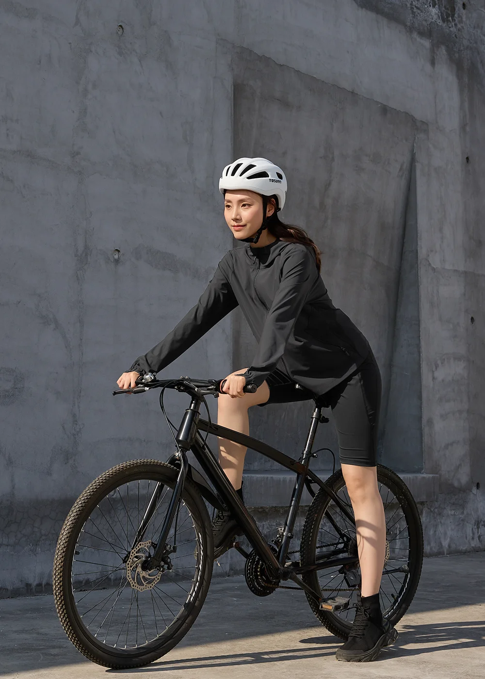 戶外機能防曬穿搭。女性模特兒穿著全黑防曬連帽外套、修身五分單車褲，配戴白色自行車安全帽，雙手緊握手把，跨騎在黑色山地單車上，背景為簡約水泥牆。