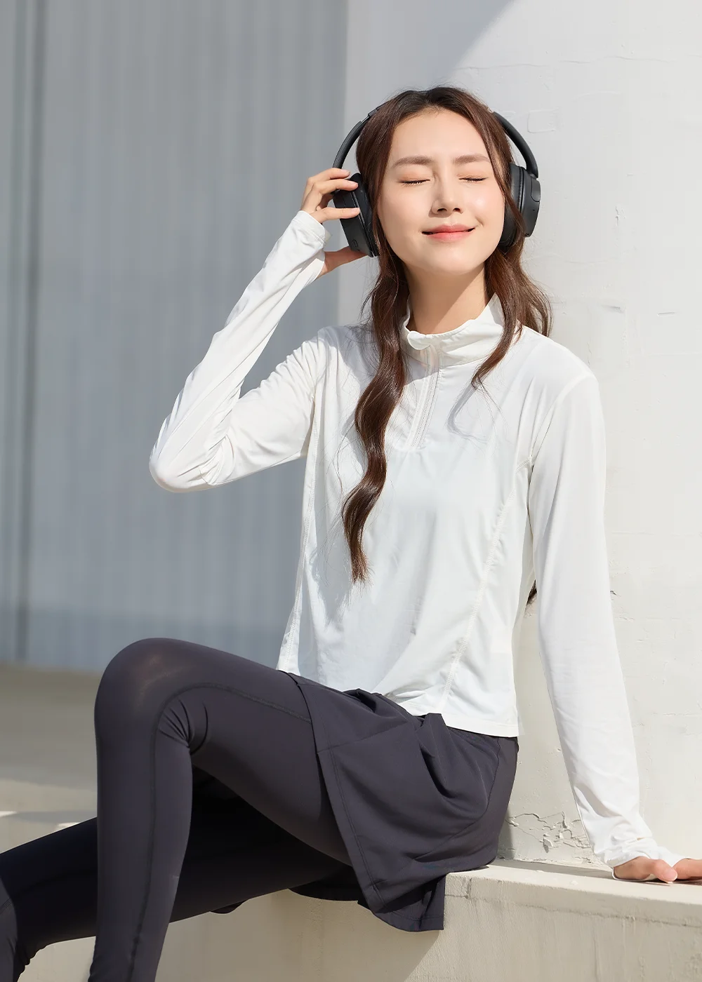 UV100 女生戶外運動防曬 OOTD：模特兒戴黑色耳機、閉眼微笑，身穿白色立領防曬長袖衫搭配黑色運動內搭褲與運動裙，在陽光下展現舒適透氣的休閒姿態。