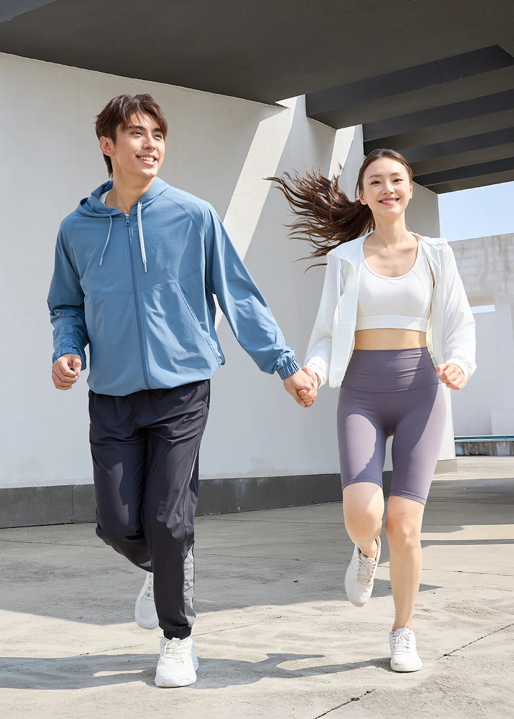 男女模特兒穿著防曬外套在戶外陽光下牽手跑步，展現外套在運動時的輕盈與舒適度。女模穿白色外套搭配灰色緊身褲，男模穿藍色外套搭配黑色運動長褲。