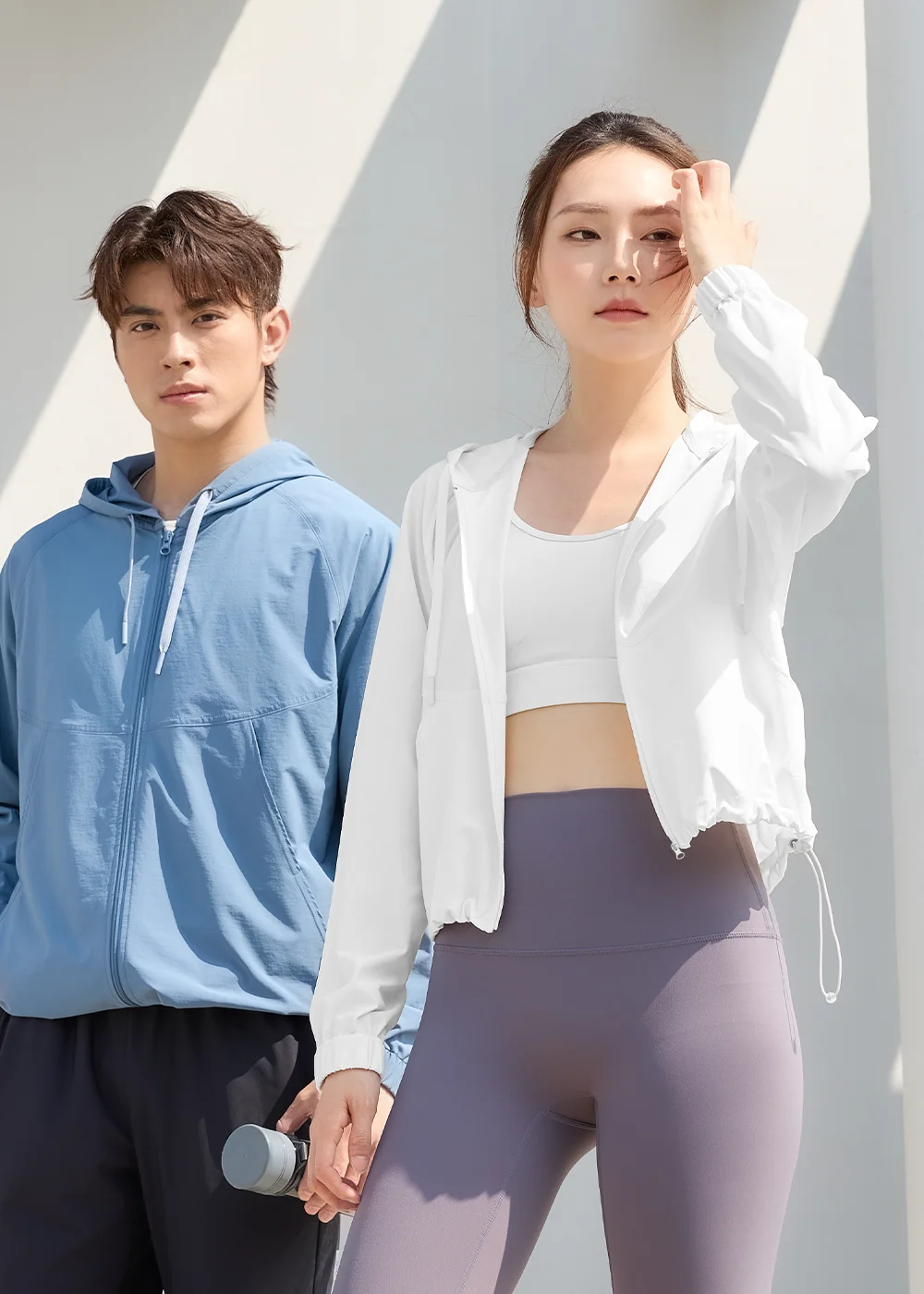 男女模特兒在陽光下穿著 Suptex 防曬外套正面合照。女模特兒身穿白色款式，手撥長髮；男模特兒身穿藍色款式，手持運動水壺，呈現簡約率性的戶外機能穿搭。