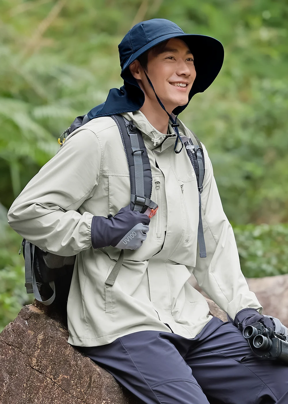 一名模特兒身穿米色戶外連帽夾克、深藍色休閒長褲，並戴著深藍色寬邊防曬遮陽帽（附護頸片）和灰色防曬手套，背著黑色背包，手拿雙筒望遠鏡坐在森林中的木頭上，微笑著看向右方。