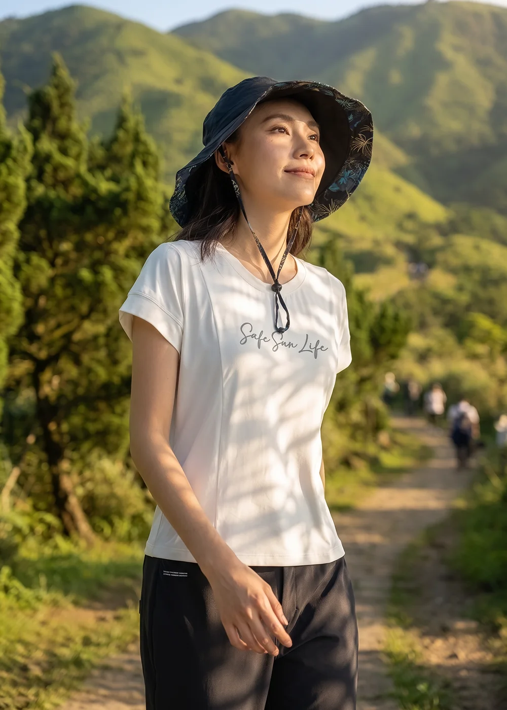 女模特兒在山林步道漫步，戴著 UV100 深色花布拼接大檐防曬漁夫帽，穿著白色機能休閒T恤與黑色長褲，展現戶外登山健行的休閒防曬風格