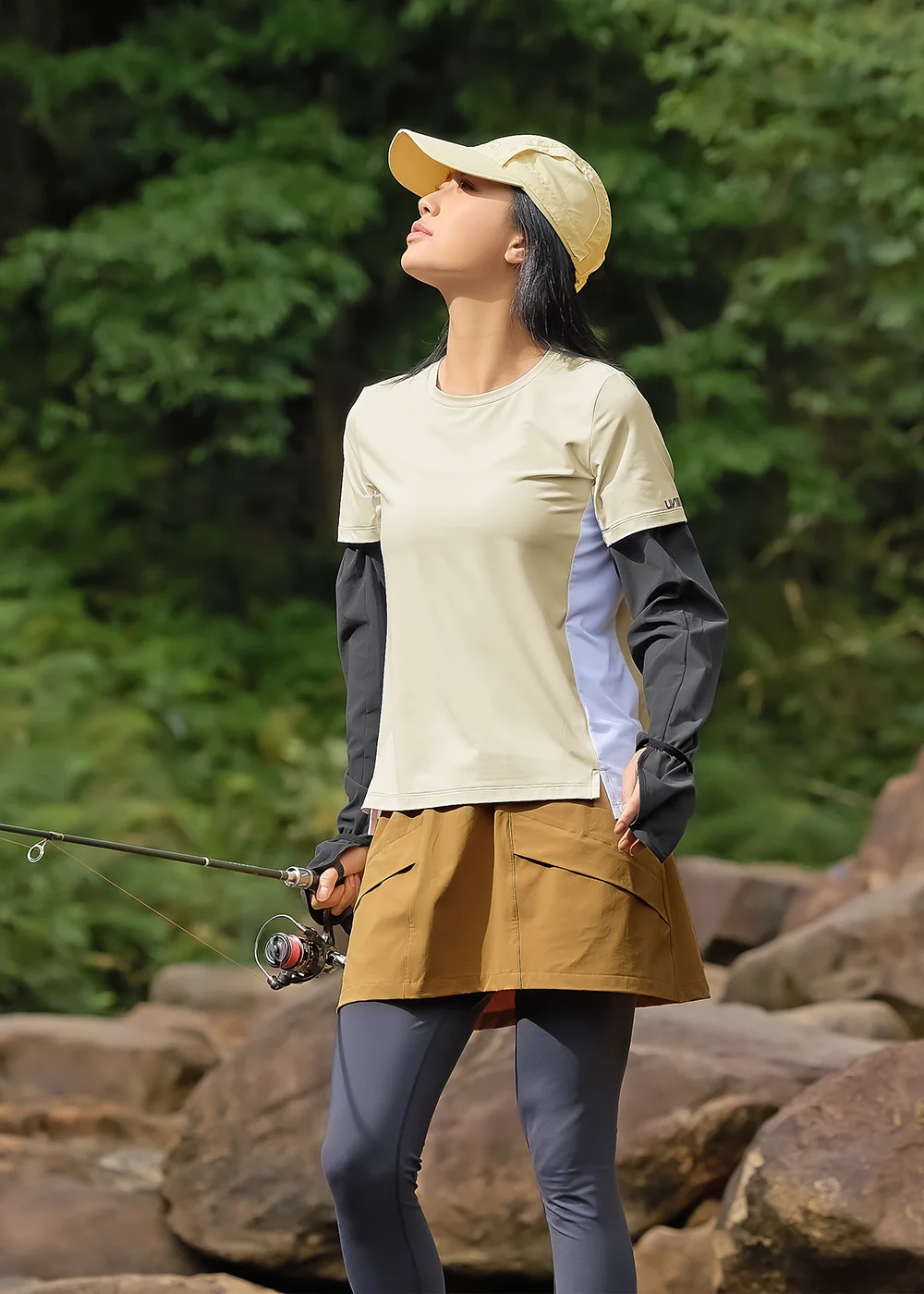 女子身著撞色機能短袖與防曬袖套，搭配卡其短裙與深色內搭褲，昂首展現俐落的野外探索穿搭。