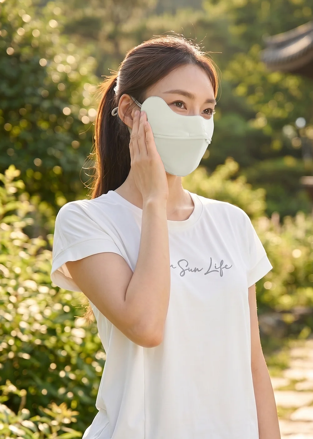 女模特兒身穿白色印有Safe Sun Life字樣的休閒短袖T恤，配戴淺色護眼角防曬涼感口罩，在陽光灑落的綠意庭園中單手輕撫臉頰，展現日常戶外運動的舒適防護