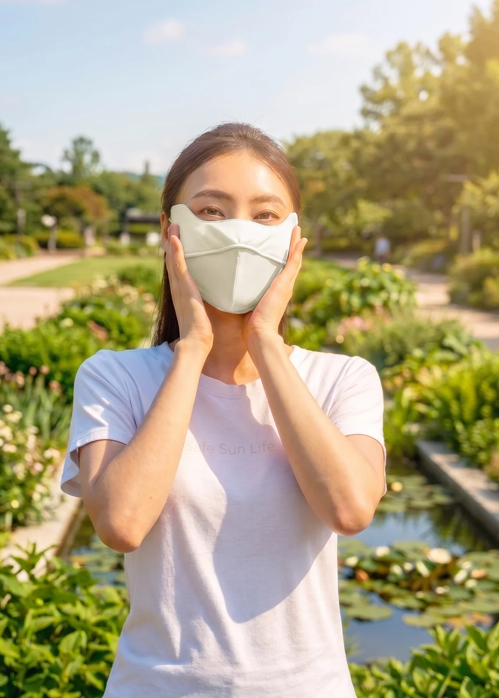 女模特兒雙手捧頰，身穿白色休閒短袖，配戴UV100淺色護眼角立體防曬口罩，背景為陽光充足的公園綠地與生態池，強調夏日安心防曬與高效紫外線阻隔
