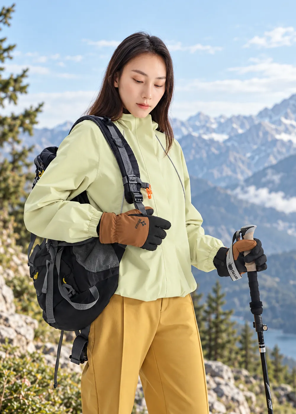 戶外登山健行情境：女性登山客身著淺綠色外套與芥黃色戶外長褲、後背登山背包，雙手佩戴棕黑拼色防寒手套並手持登山杖，展現科技羊毛手套於山林健行時的防護與活動性。