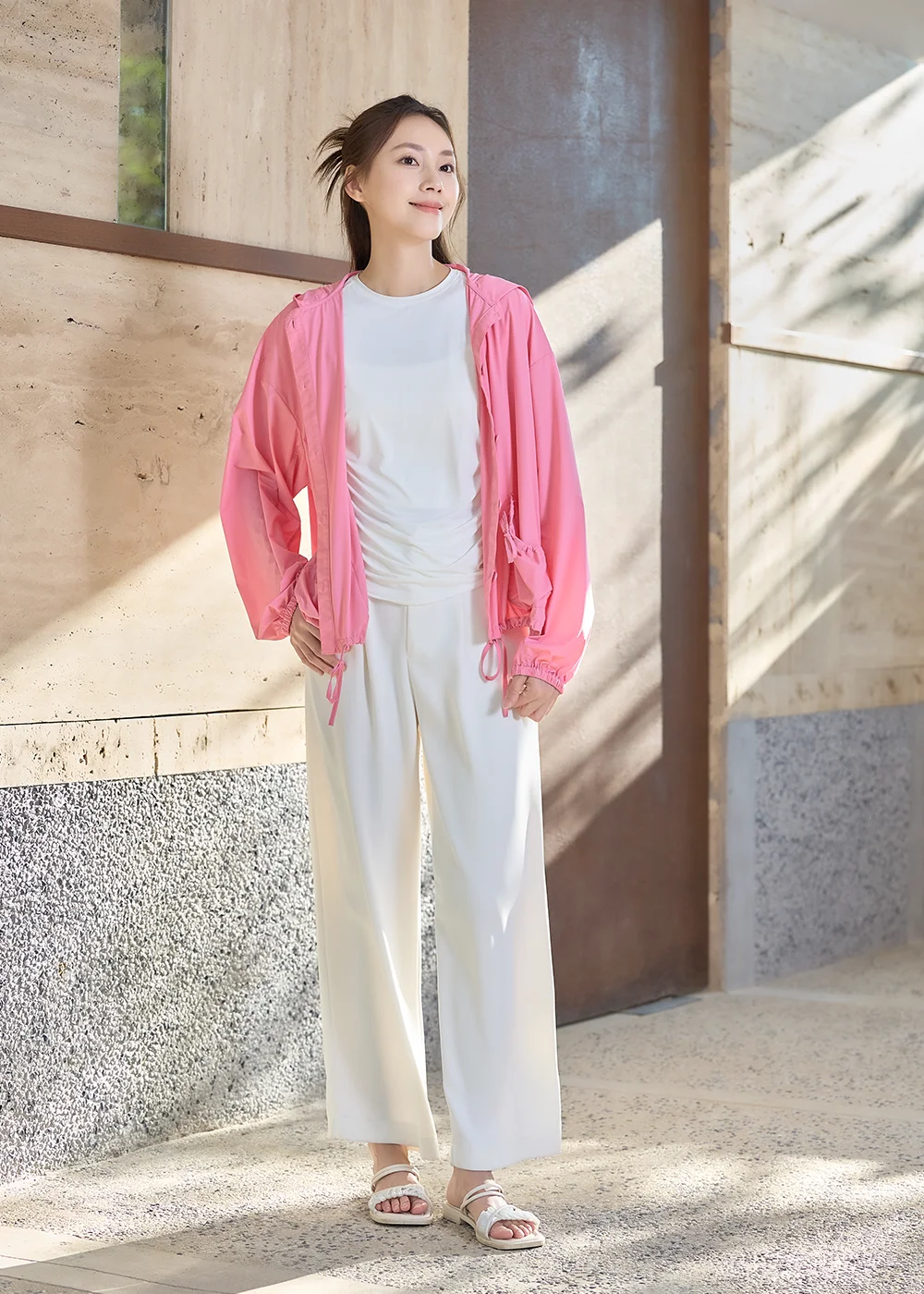 UV100 女生夏天涼感防曬衣：模特兒全身示範粉紅色連帽防曬外套、內搭白色 T 恤與白色抽繩休閒長褲，搭配白色涼鞋的清爽遮陽穿搭。